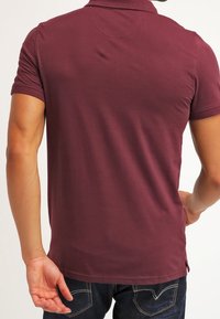 Polo bordeaux à manches courtes, col classique et fentes latérales. Tissu doux et lisse avec une coupe ajustée. Porté avec un jean bleu foncé.