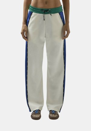 Persona che indossa pantaloni della tuta bianchi con fascia elastica verde in vita, pannelli laterali blu, coulisse nere e scarpe marroni con lacci blu.