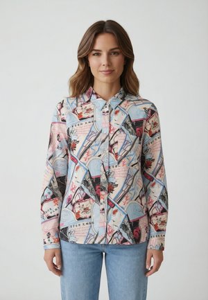 Donna che indossa una camicetta con collo e bottoni, caratterizzata da una stampa astratta e floreale colorata, abbinata a jeans azzurri, in piedi davanti a uno sfondo semplice.