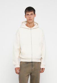 White zip-up hoodie with a drawstring hood, front pocket, and ribbed cuffs, paired with tan pants. Smooth fabric, relaxed fit.