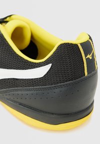 Pantof de sport negru cu superior din plasă, accente galbene și un logo alb. Prezintă un toc texturat și detalii perforate pe lateral.