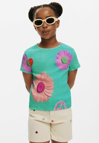 Tyrkysová krátkosrstá košeľa s veľkými vyšívanými kvetmi v ružovej, fialovej a červenej farbe, skombinovaná s béžovými šortkami zdobenými malými kvetinovými vzormi.