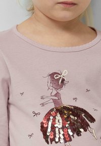 Rosa Langarmshirt mit einer Illustration eines Mädchens in einem braunen Kleid, verziert mit Pailletten in Rottönen und Rosa, mit einer Schleifen-Akzent.