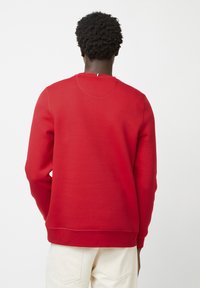 Sweatshirt rouge en tissu doux, avec un col rond et des poignets côtelés. Le design au dos comprend un petit détail au niveau du col.