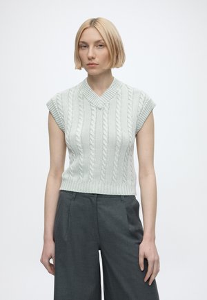Kvinde med kort blond hår iført en ærmeløs lysegrå kabelstrikket sweater og mørkegrå bukser med vide ben, stående foran en ensfarvet baggrund.