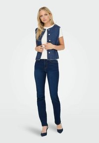 Denimová bezrukávová vesta s pěti knoflíky, nošená přes bílý tričko, spárovaná s tmavě modrými skinny džínami a tmavě modrými podpatky.