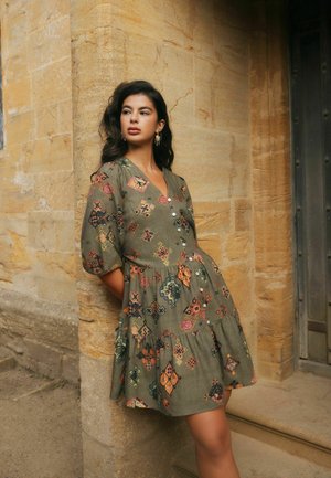 Woman leans against textured stone wall wearing olive green dress with colorful geometric patterns and buttons, standing near wooden door.