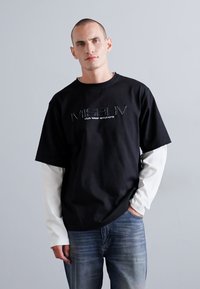 Svart kortermet t-skjorte med "MISBHV"-logo, lagdelt over en hvit langermet topp, sammen med blå denimjeans. Enkel design og rene linjer.