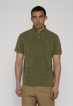 Mann med olivengrønn kortermet poloshirt og lysebrune bukser, står mot ensfarget lysegrå bakgrunn.