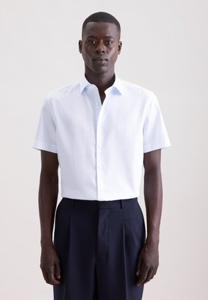 Schwarzer Mann steht, trägt ein weißes Kurzarm-Hemd mit Knöpfen und dunkle hochgeschnittene Hosen, schaut nach vorne vor einem einfarbigen Hintergrund.