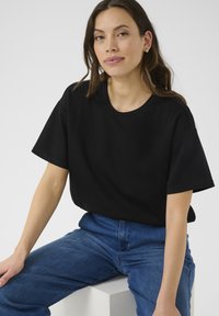 T-shirt nera oversize con scollatura rotonda e maniche corte, realizzata in un tessuto morbido. Abbinata a jeans in denim blu, seduta su uno sfondo neutro.