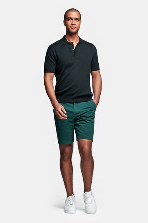 Schwarzes Poloshirt mit kurzen Ärmeln und Kragen, kombiniert mit grünen Shorts und weißen Sneakers. Der Look ist lässig und monochrom.