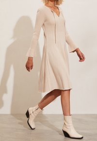 Robe tricotée à côtes, longueur genou, en beige clair avec un décolleté en V, des manches longues et des poignets évasés. Associée à des bottines blanches à talons.