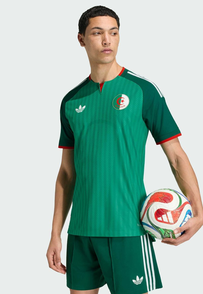 Jeune homme en tenue de football algérienne verte tenant un ballon de football Adidas coloré sur un fond uni.