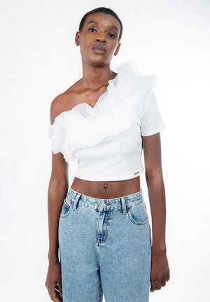 Kvinde iført hvid one-shoulder crop top med flæser og lyseblå højtaljede jeans, stående mod en ensfarvet hvid baggrund.