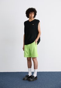 Pull en maille sans manches noir, short vert fluo avec logo, chaussettes blanches et chaussures à plateforme noires, présentés sur un mannequin masculin sur fond neutre.