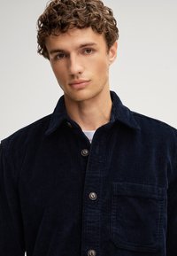 Camicia di velluto a coste blu navy con chiusura a bottoni, colletto a punta, taschino sul petto e tessuto testurizzato, dotata di bottoni in tonalità argentata.