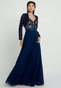 Nocna suknia w kolorze granatowym z przeźroczystą długą narzutą, koronkowym zwieńczeniem oraz zwiewną spódnicą. Posiada dekolt w kształcie litery V i ozdobny pas.