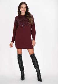 Burgunderfarbendes Strickkleid mit Rundhalsausschnitt und schimmernden Perlenverzierungen. Getragen mit schwarzen Lederstiefeln bis zum Oberschenkel.