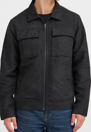 Homme portant une veste noire zippée avec deux poches à rabat sur la poitrine et un jean bleu sur fond uni.