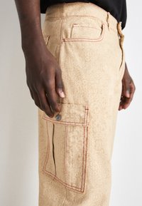 Beige byxor med ett subtilt mönster av logotyper över hela ytan, kontrasterande röda sömmar och en framträdande ficka med tryckknappsdetalj.