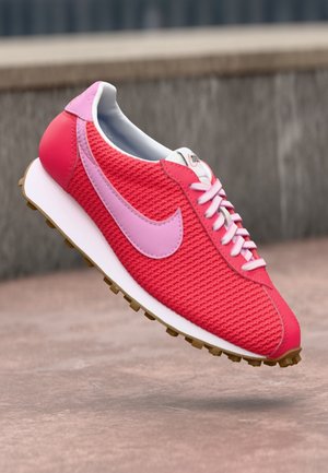 Czerwony i różowy but do biegania Nike z teksturowaną cholewką z tkaniny i białymi sznurówkami, pokazany pod kątem na tle zewnętrznego kamienia.