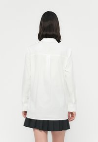 Camisa branca de botões com mangas longas, apresentando um yoke nas costas e uma bainha reta, combinada com uma saia preta plissada.