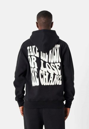Zwarte hoodie van katoen met een voorzak. Beschikt over grote witte grafische tekst op de achterkant: "NEEM GEEN RISICO OF VERLOPEN DE KANS."
