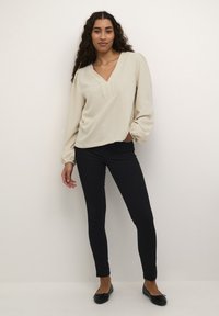 Beige Bluse mit langen Ärmeln, V-Ausschnitt und strukturierter Oberfläche, kombiniert mit schwarzen, figurbetonten Hosen und schwarzen flachen Schuhen, vor einem neutralen Hintergrund stehend.