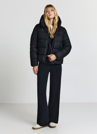 Giubbotto imbottito nero con cappuccio, caratterizzato da un design trapuntato e zipper frontale, indossato sopra una maglietta nera e abbinato a pantaloni nero svasati.