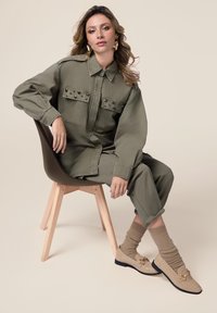 Camicia oversize verde oliva con dettagli in borchie, pantaloni abbinati, mocassini beige con accenti dorati e calzini a coste beige. Sedia elegante in legno.
