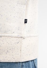 Gros plan sur l'ourlet d'un sweat-shirt crème tacheté avec un détail de poignet côtelé et une petite étiquette de marque noire, porté sur un jean en denim bleu.