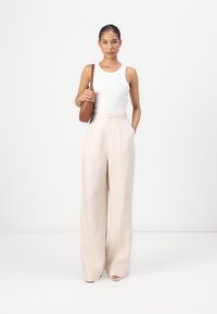 Top sin mangas blanco con cuello alto, combinado con pantalones de pierna ancha beige claro. Lleva un bolso de hombro marrón redondo. Texturas suaves.
