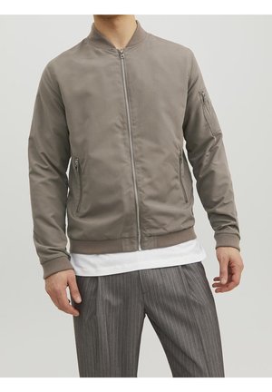Homme portant une veste bomber taupe avec des poches zippées et un pantalon gris à fines rayures, debout devant un fond uni.