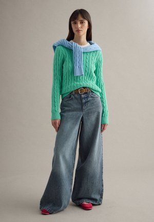 Young woman wearing a green cable-knit sweater, wide-leg blue jeans, pink shoes, and a light blue sweater draped over her shoulders.