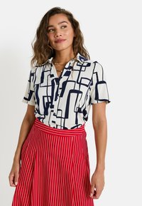 Blouse à manches courtes en crème avec imprimé géométrique abstrait bleu marine, associé à une jupe rouge à rayures. La blouse a une fermeture à boutons et un col.