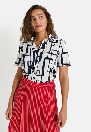Blusa de manga corta en crema con estampado geométrico abstracto en azul marino, combinada con una falda roja a rayas. La blusa tiene cierre de botones y cuello.