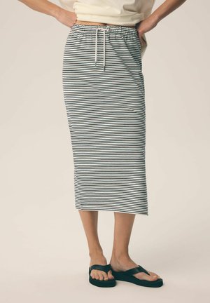Kobieta ubrana w midi spódnicę w białe i czarne paski z troczkiem w talii oraz czarne klapki na palce, stoi z rękami na biodrach.