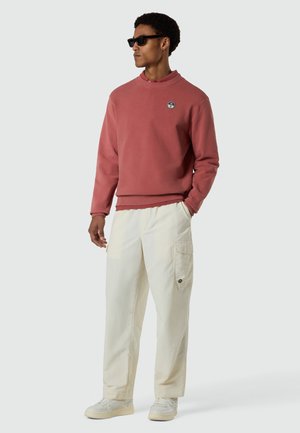 Uomo con occhiali da sole, felpa color rosa cipria con logo, pantaloni cargo color crema e sneakers, in piedi con una mano in tasca, guardando a sinistra.