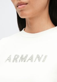 Λευκό μπλουζάκι με τη λέξη "ARMANI" σε μεγάλα, ασημένια, γκλιτερ γράμματα. Λείο ύφασμα με λαιμόκοψη τύπου crew, κοντινή όψη.