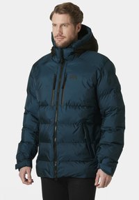 Chaqueta acolchada de color azul oscuro con un acabado brillante, capucha negra, cierre frontal con cremallera y dos bolsillos en el pecho con cremallera. Presenta puños elásticos y un diseño acolchado.