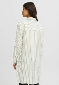 Tunic leggera color crema con strisce verticali nere, maniche lunghe e colletto piccolo; presenta una vestibilità ampia e una texture morbida.