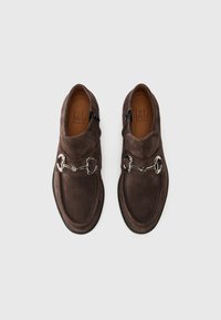 Zapatos de ante marrón con una textura suave, que presentan un detalle de cadena plateada en la parte superior, puntera redondeada y costuras de suela negra contrastantes.