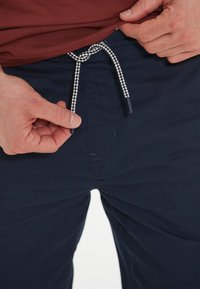 Navyblaue Hosen mit einem Kordelzugbund, der mit einer schwarz-weißen geflochtenen Schnur versehen ist. Sichtbare Nähte und ein glattes, strukturiertes Gewebe.