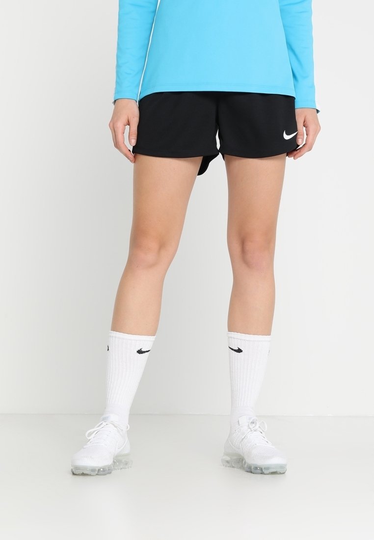 Personne portant un short Nike noir, des chaussettes Nike blanches, des chaussures de sport blanches et un haut à manches longues bleu, debout sur un sol blanc.