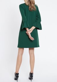 Grön klänning med långa, utsvängda ärmar och rak skärning, tillverkad av slätt tyg. Bärs med svarta högklackade skor.