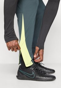 Nike kalhoty ve tmavě zelené a černé s jasně žlutým bočním panelem. Tkanina je hladká a přiléhavá, doplněná o černé sportovní boty.