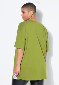 Camiseta de algodón verde de gran tamaño con mangas cortas, cuello redondo y detalle de etiqueta lateral. Textura suave y diseño simple.