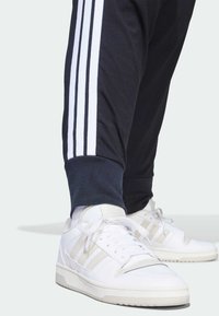 Črne trenirke s tremi belimi črtami ob straneh, združene z belimi supergami z svetlo sivimi poudarki in teksturiranimi detajli.