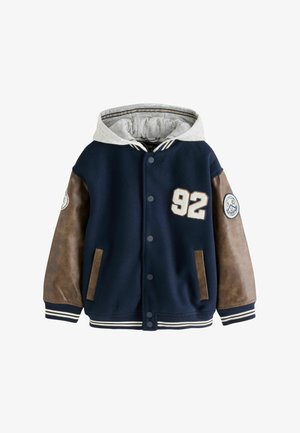 Smilende tumling iført en marineblå varsityjakke med læderærmer, blå jeans, hvide sneakers, stående mod en ensfarvet baggrund.
